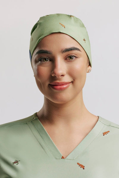 Frontal portrait of a smiling female healthcare worker in sage green scrubs wearing a matching sage green tie-back scrub cap, both printed with small platypus illustrations.