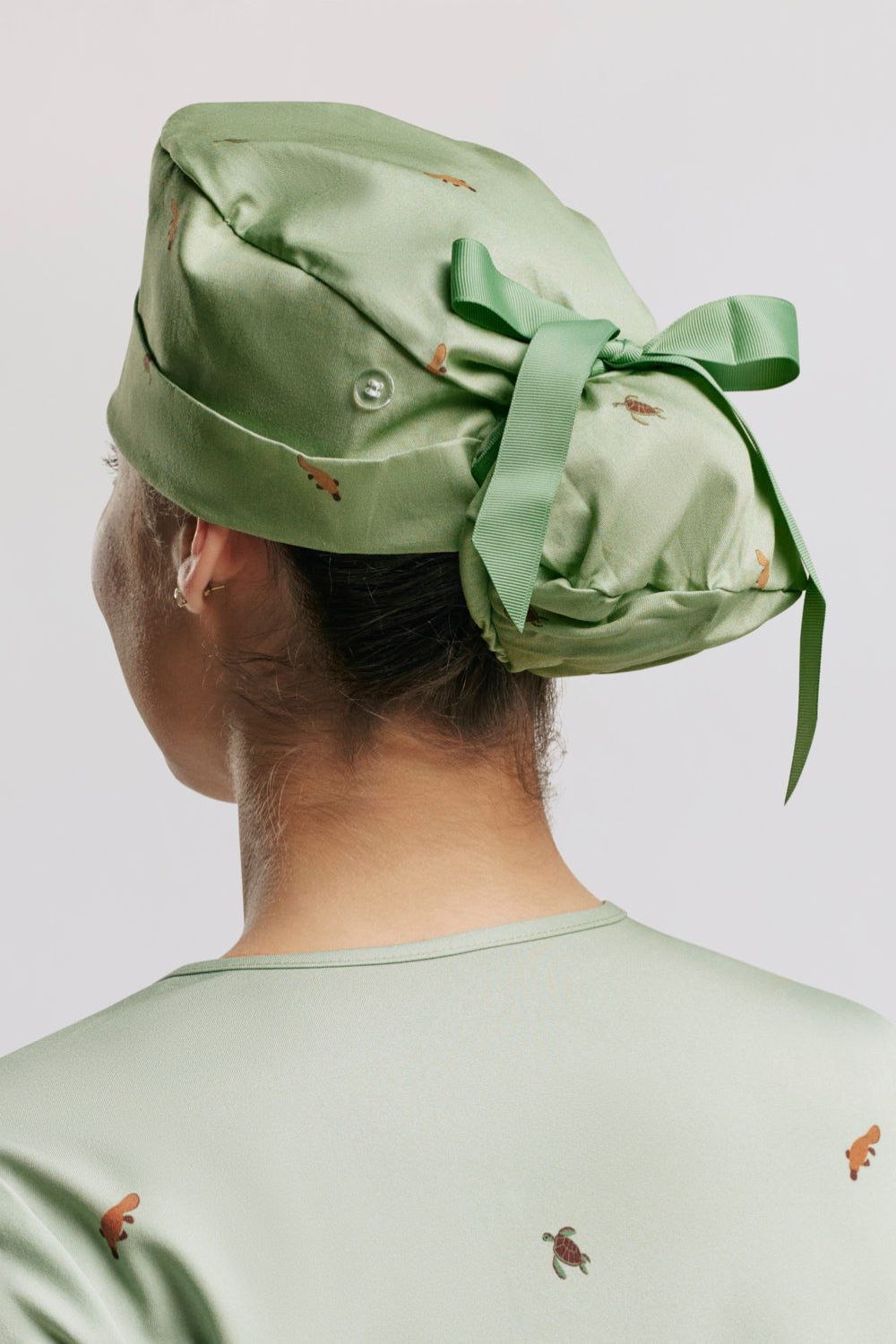 Direct rear view showing a sage green platypus-print scrub cap secured over hair in a bun with a matching sage green ribbon bow tie.