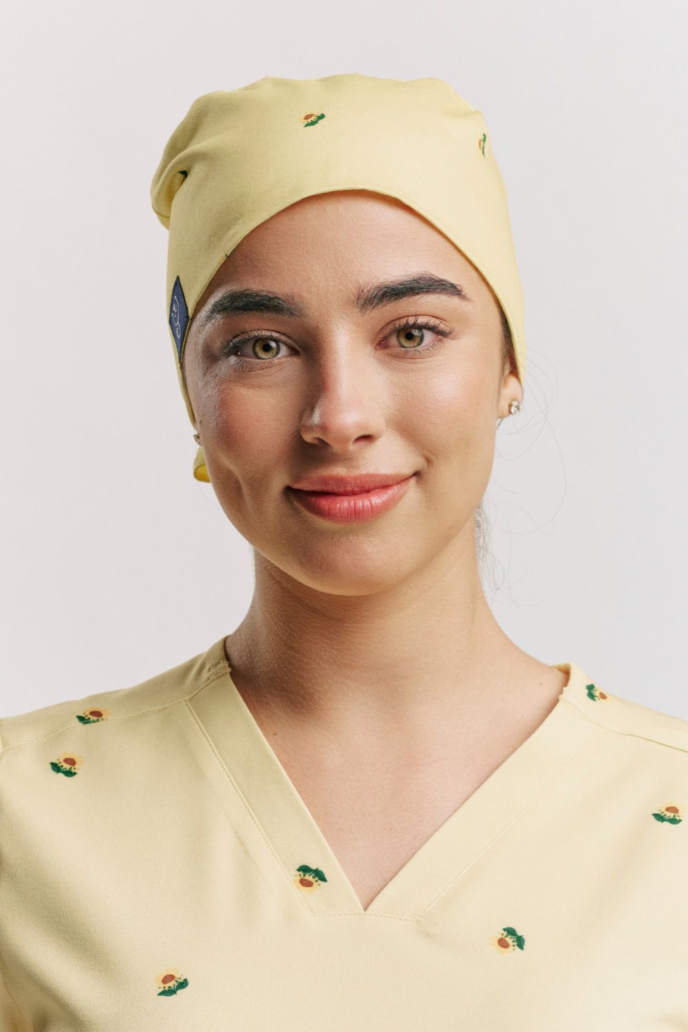 Frontal portrait of a smiling female healthcare worker in pale yellow scrubs wearing a matching pale yellow tie-back scrub cap, both printed with tiny orange and green plant/flower motifs.