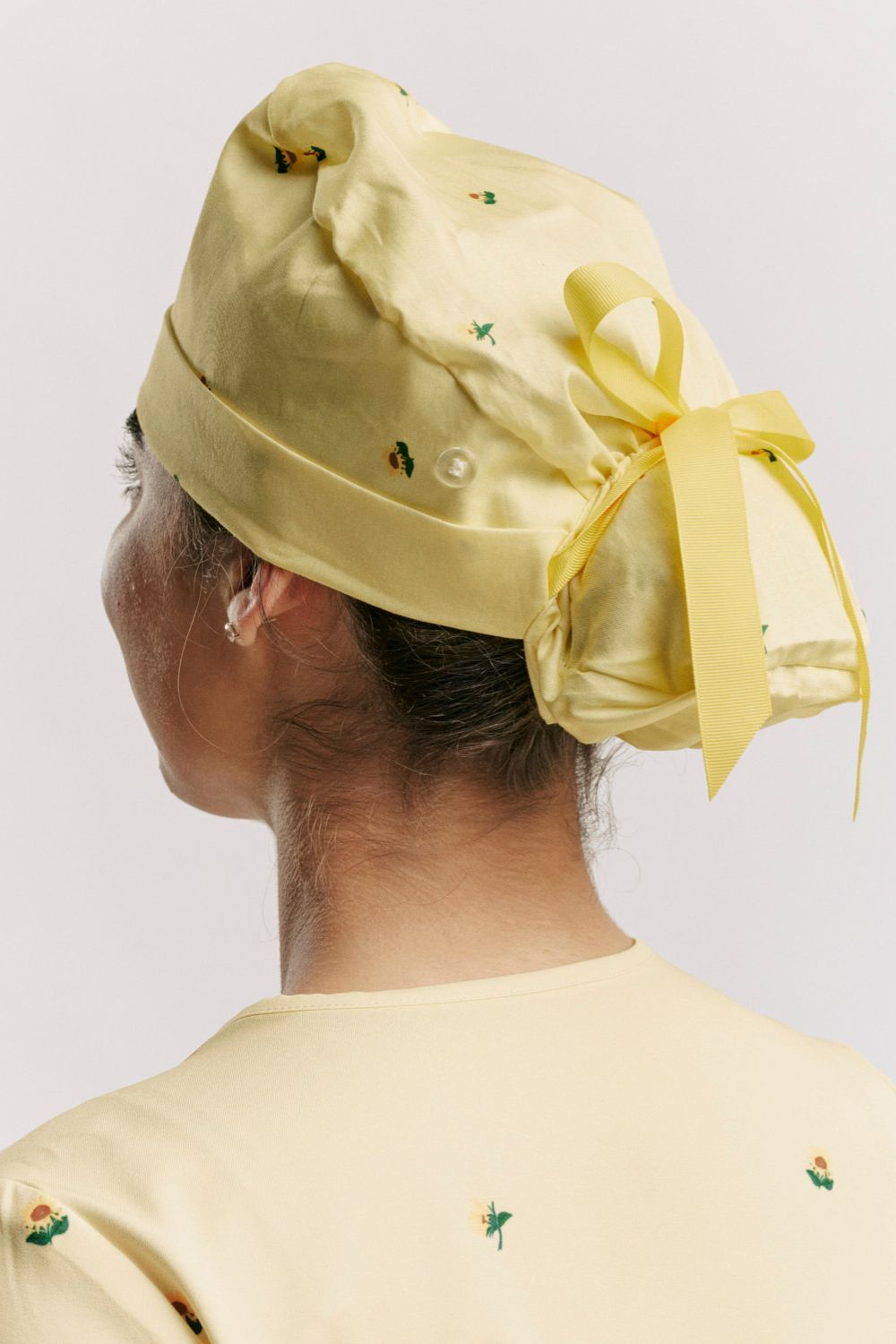 Direct rear view showing a pale yellow print scrub cap secured over hair in a ponytail pouch with a matching pale yellow ribbon bow tie.