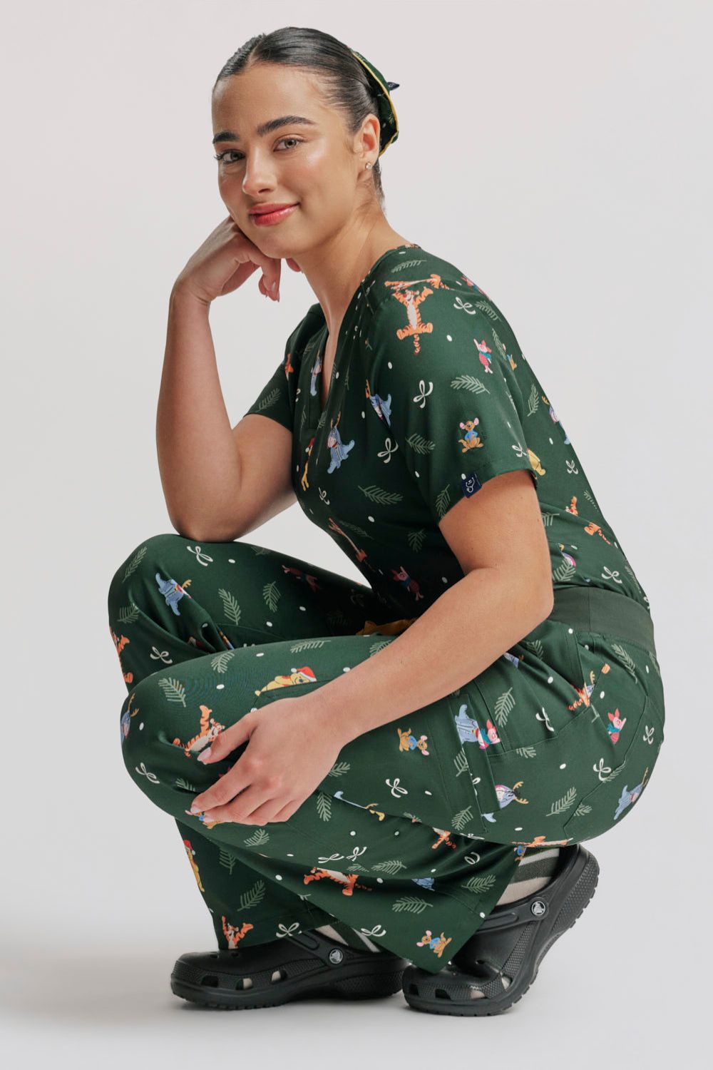 A doctor squatting/crouching in the dark forest green Winnie the Pooh scrub top and matching pants, styled with black Clogs.