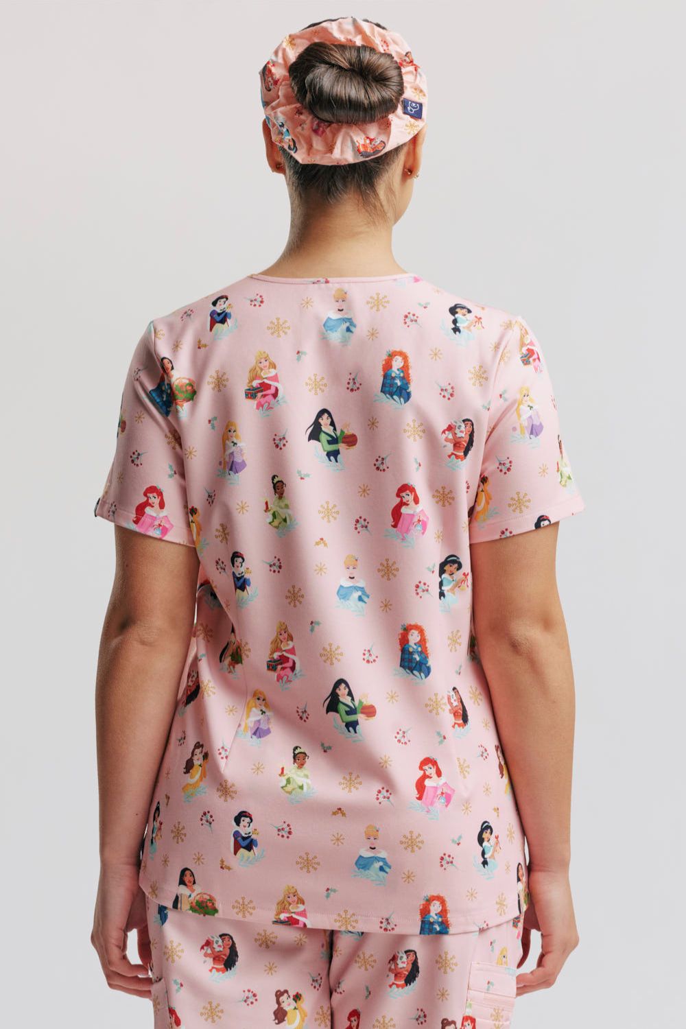Direct rear view of a female model wearing the light pink Disney Princess Christmas scrub top, matching scrub pants, and a patterned hair scrunchie/cap.