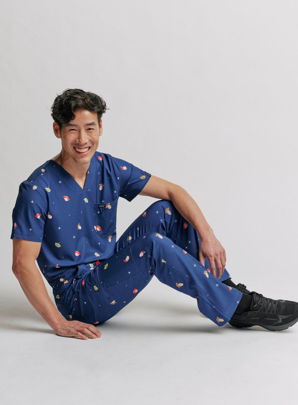 A healthcare worker leaning back seated on the floor, wearing the dark blue Men's Toy Story Christmas scrub top and matching scrub pants set.