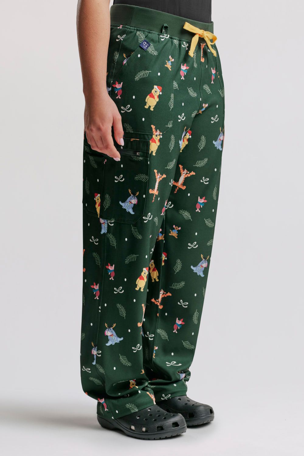 Side profile of a healthcare worker wearing the dark forest green Winnie the Pooh scrub pants, highlighting side pocket detail, leg fit, and black Clogs.