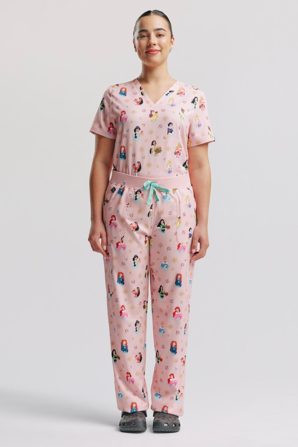 A healthcare worker standing in the matching light pink Disney Princess Christmas V-neck scrub top and cargo scrub pants set.