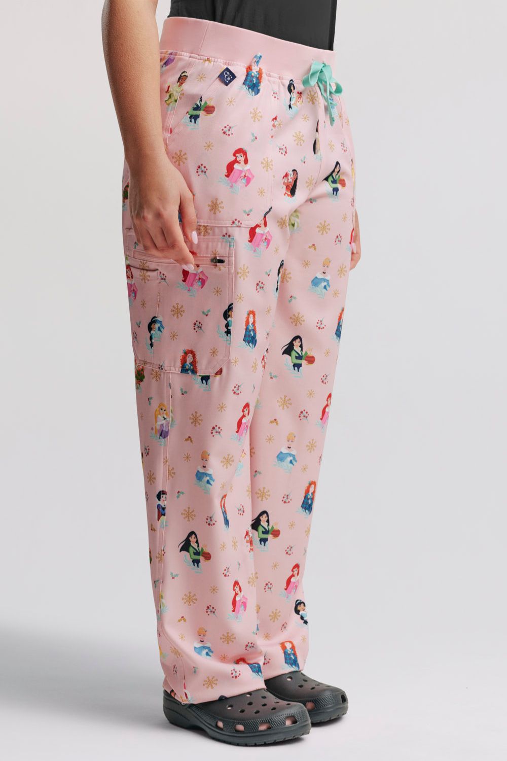 Side profile of a nurse wearing the light pink Disney Princess Christmas cargo scrub pants, highlighting the side pocket placement and leg fit, styled with dark grey Clogs.