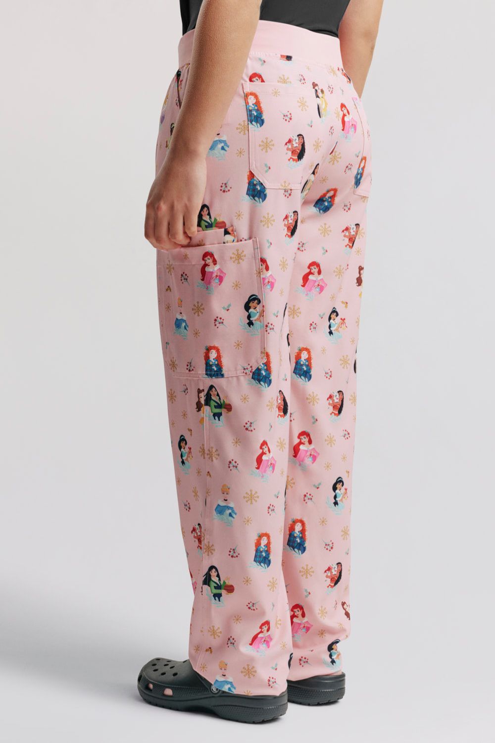 Rear three-quarter view showing the elastic waistband fit, leg drape, and pattern layout on the back of the light pink Disney Princess Christmas scrub pants, paired with dark grey Clogs.