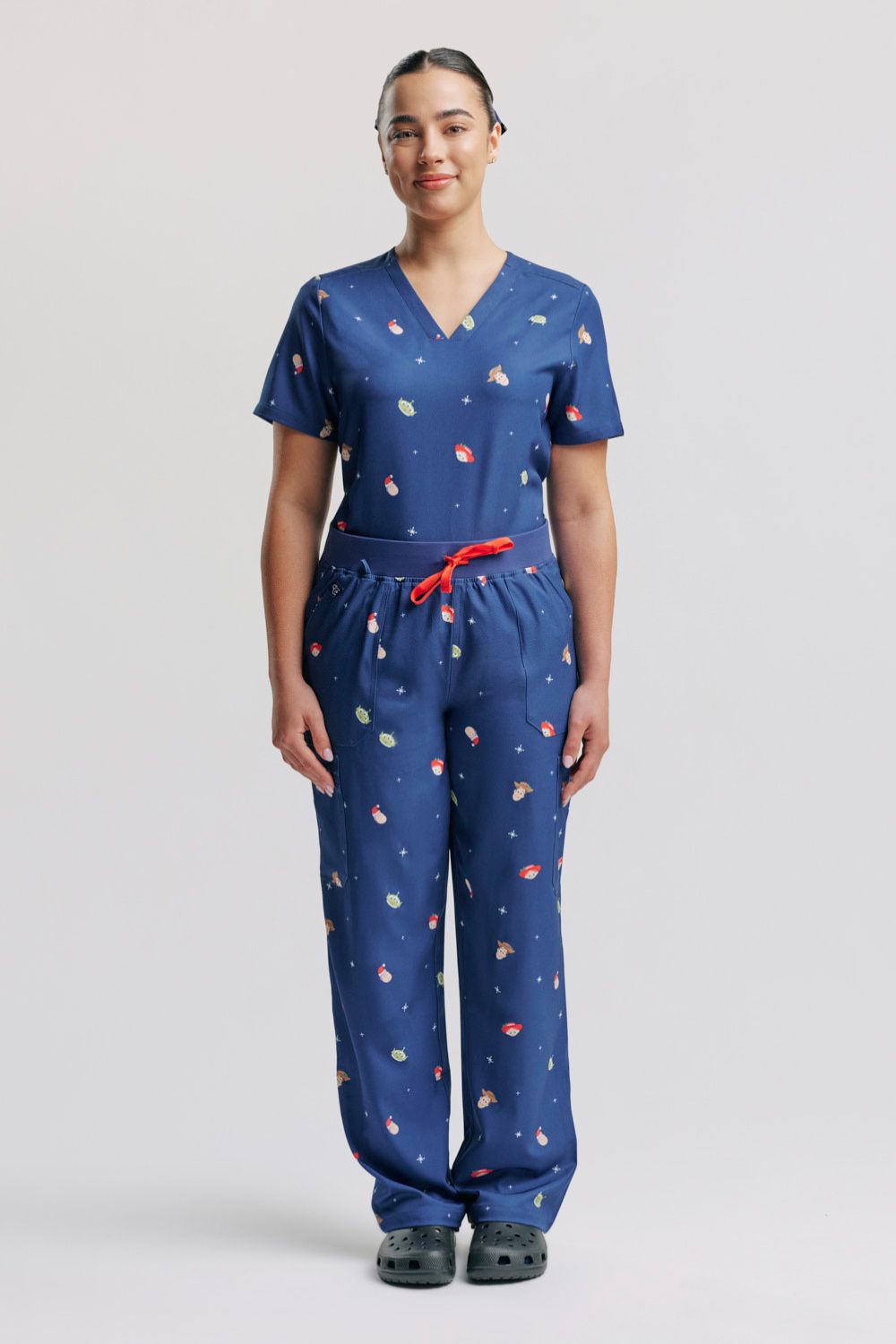 A female healthcare worker standing in the matching dark blue Disney & Pixar Toy Story Christmas V-neck scrub top and cargo scrub pants set.