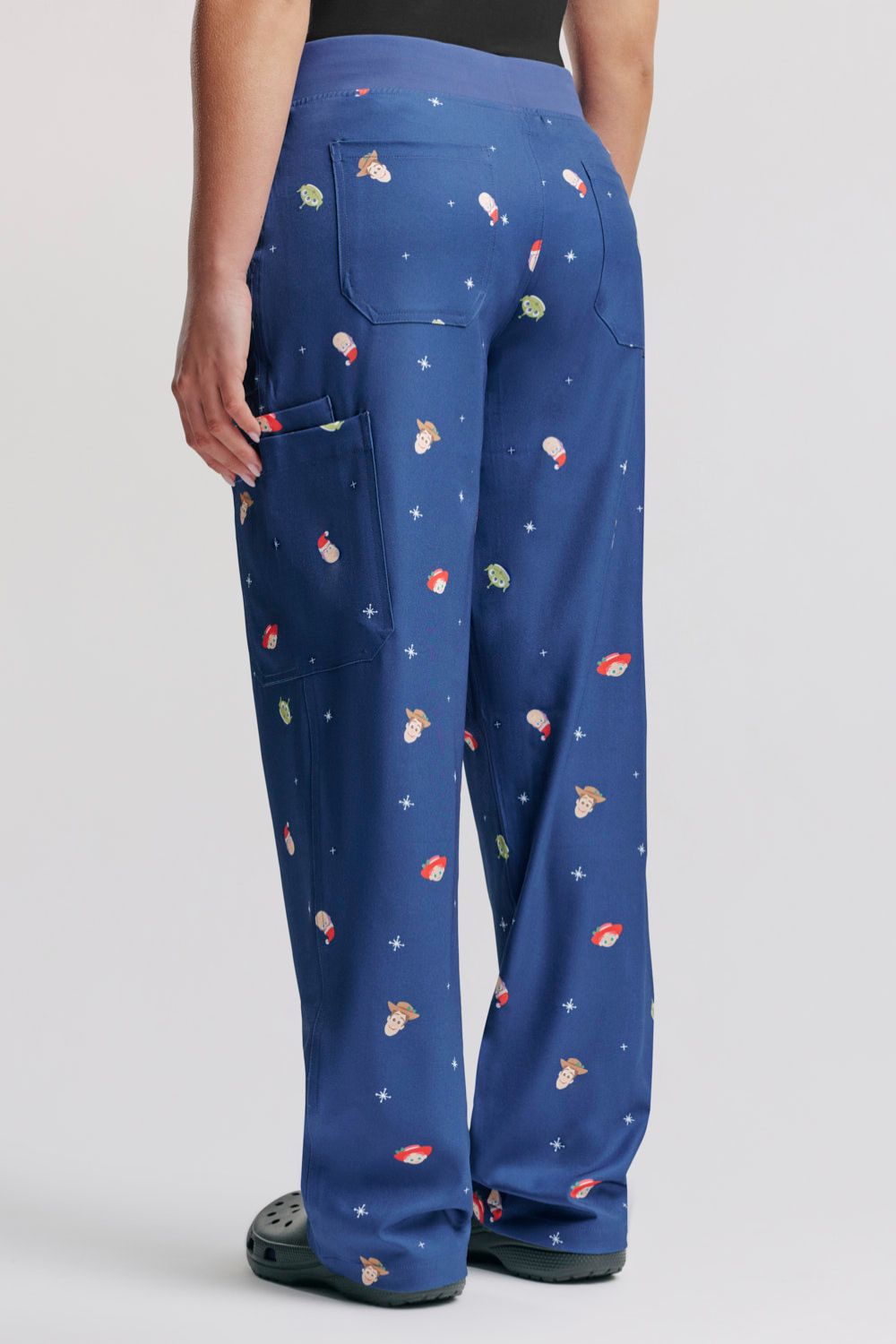 Rear three-quarter view showing the back patch pocket placement, leg drape, and holiday pattern layout on the back of the dark blue Toy Story scrub pants, paired with dark grey Clogs.