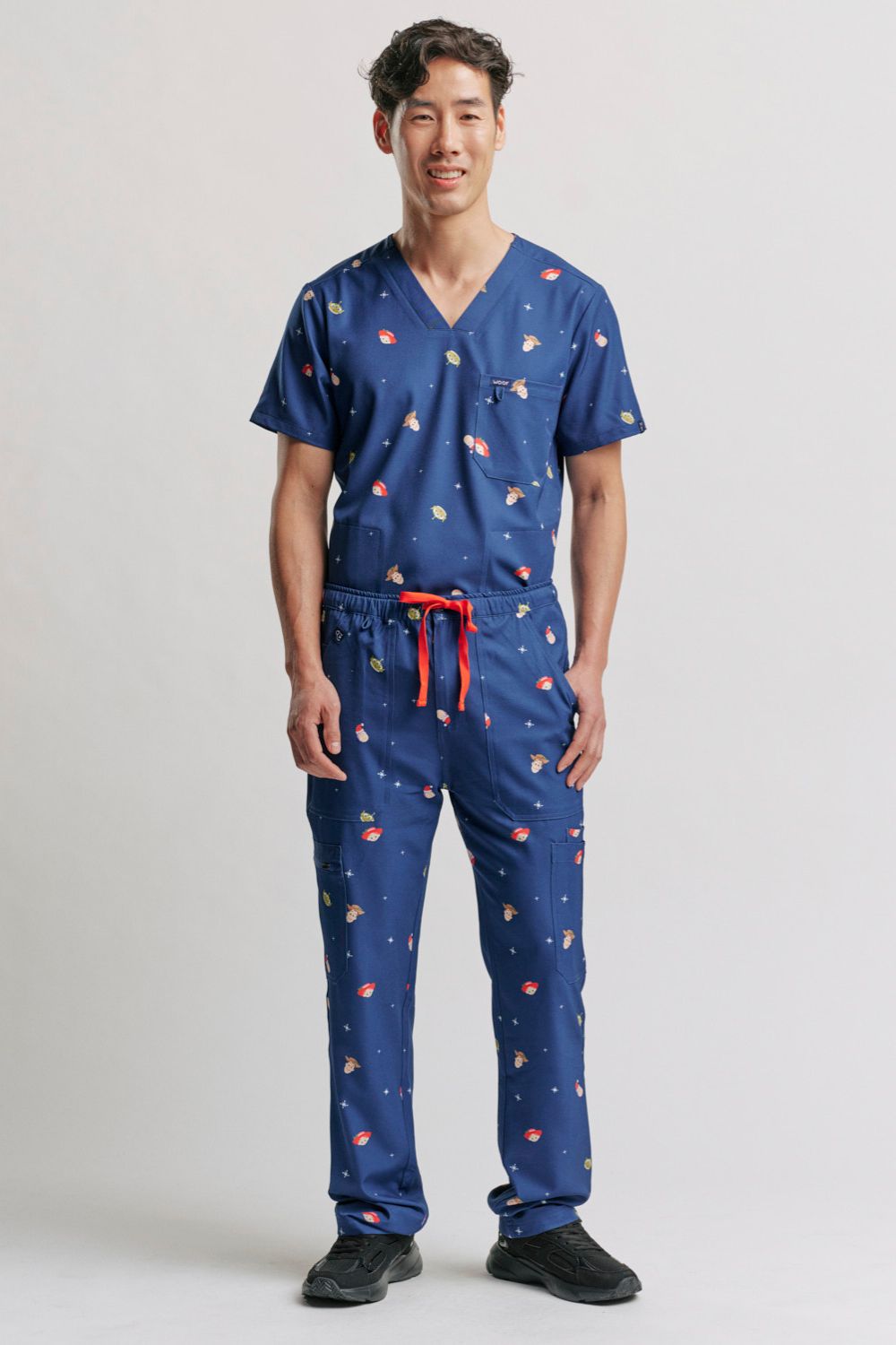 A doctor standing with hands in pockets, wearing the full dark blue Men's Toy Story Christmas V-neck scrub top and cargo pants set with black athletic shoes.