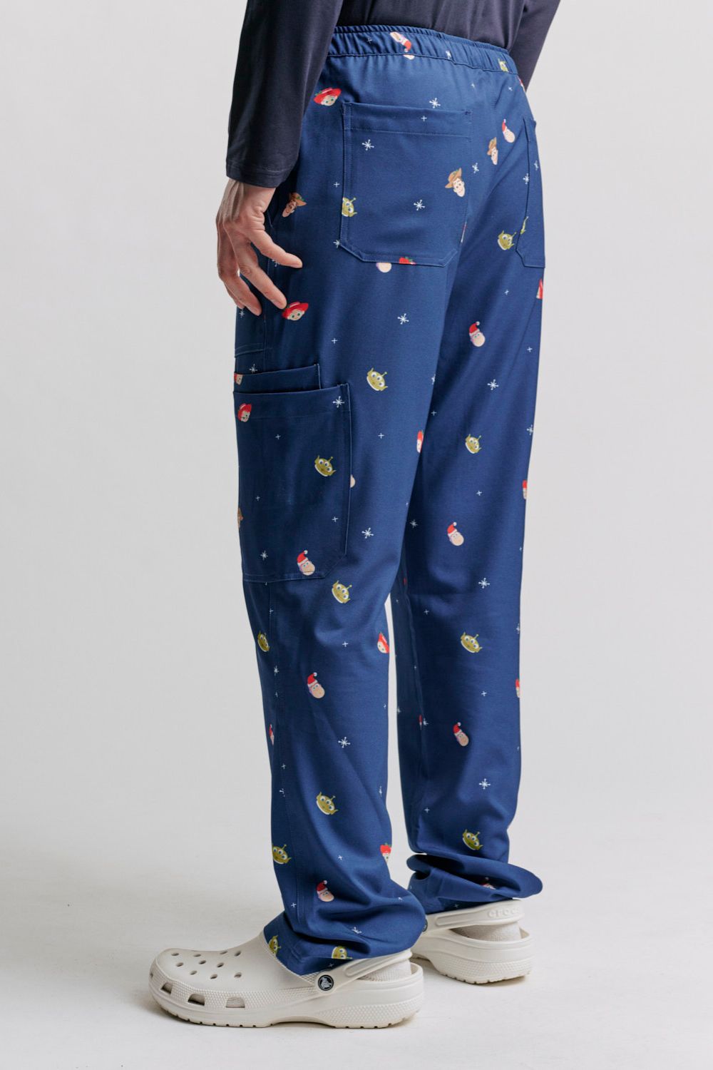 Rear three-quarter studio view showing the back patch pocket placement, side cargo pocket detail, and leg fit on the dark blue Men's Toy Story Christmas scrub pants, styled with white Clogs.