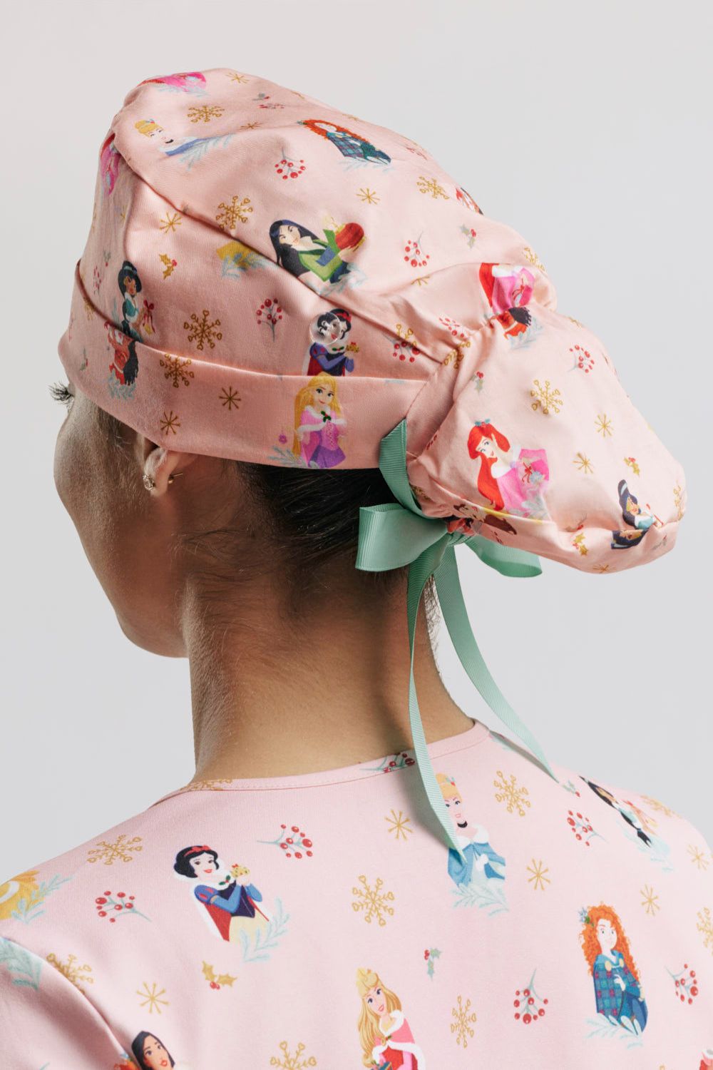 The back profile of the light pink Disney Princess Christmas scrub cap, showing the elasticized ponytail pouch secured with a mint green ribbon bow, along with the holiday character print featuring Mulan, Ariel, Rapunzel, Snow White, and Merida alongside golden snowflakes and festive holly accents.