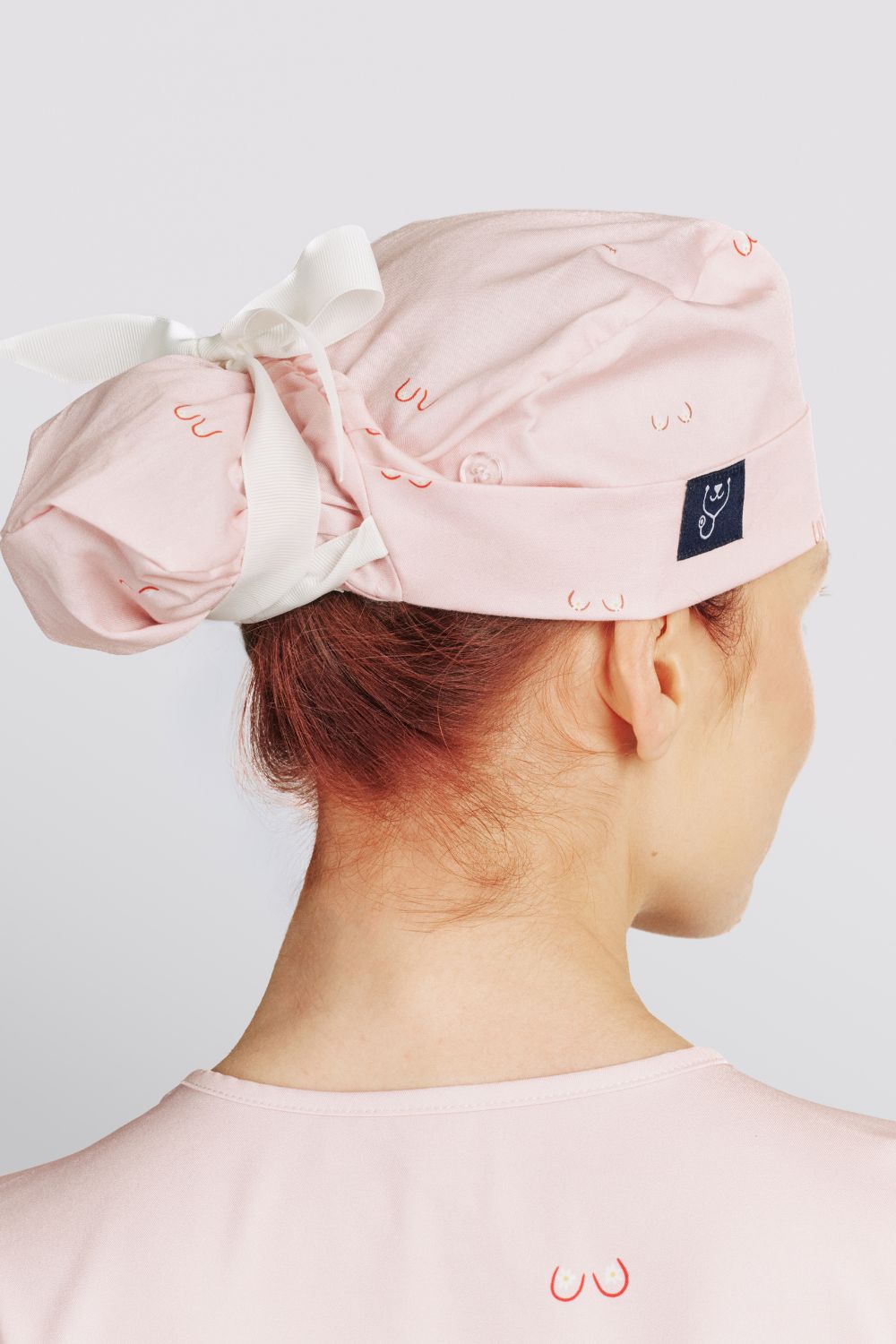 Back view of a veterinarian wearing a light pink scrub cap with line art small boobs print tied with a white ribbon.