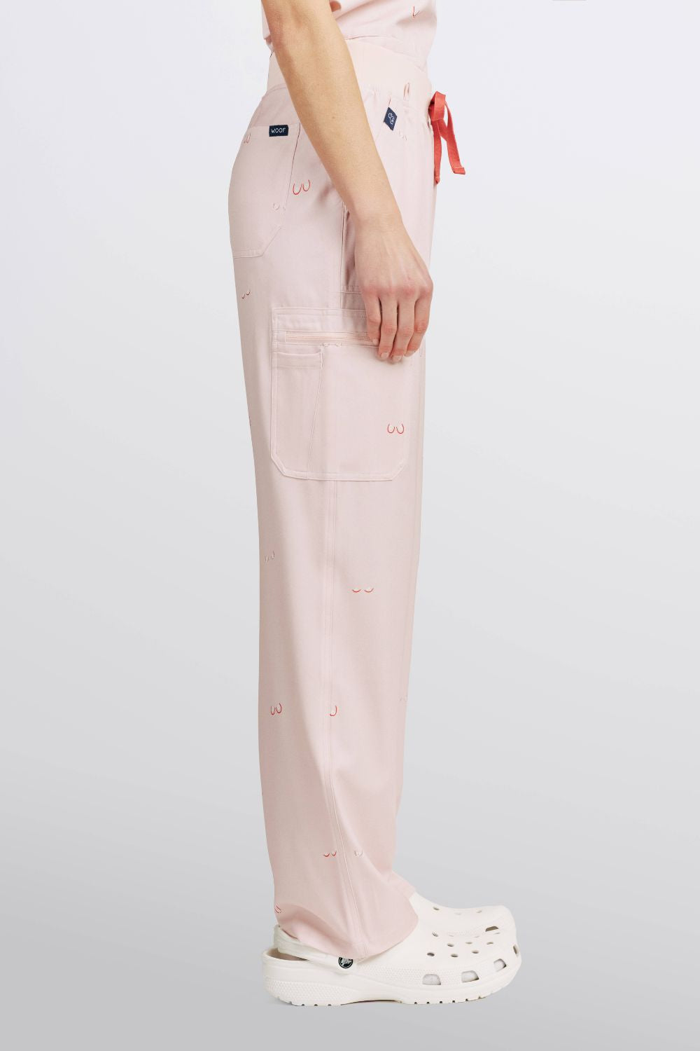 Side profile of a vet nurse wearing light pink scrub pants with line art small boobs print and white clogs.