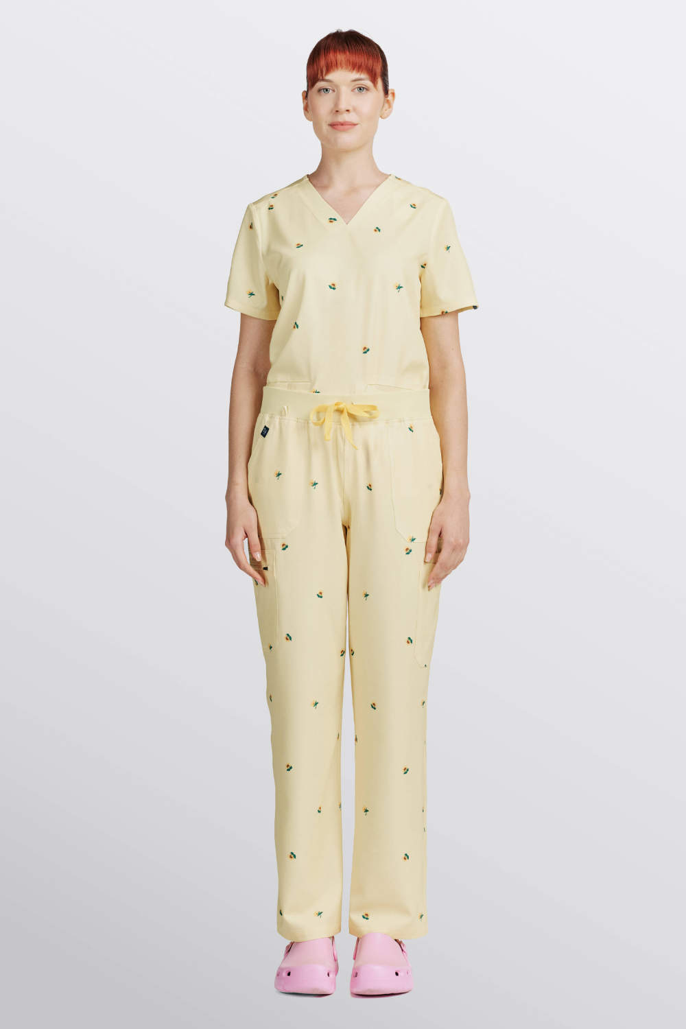 Doctor standing in full light yellow scrub set with sunflower print and pink clogs.