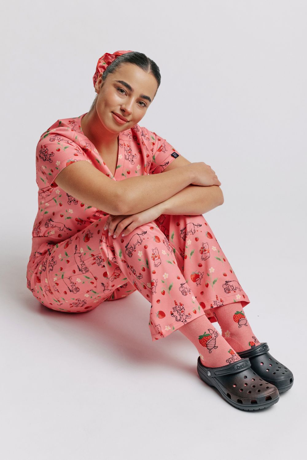 Nurse sitting on the floor in light pink scrub pants with Winnie the Pooh strawberry print and matching top.
