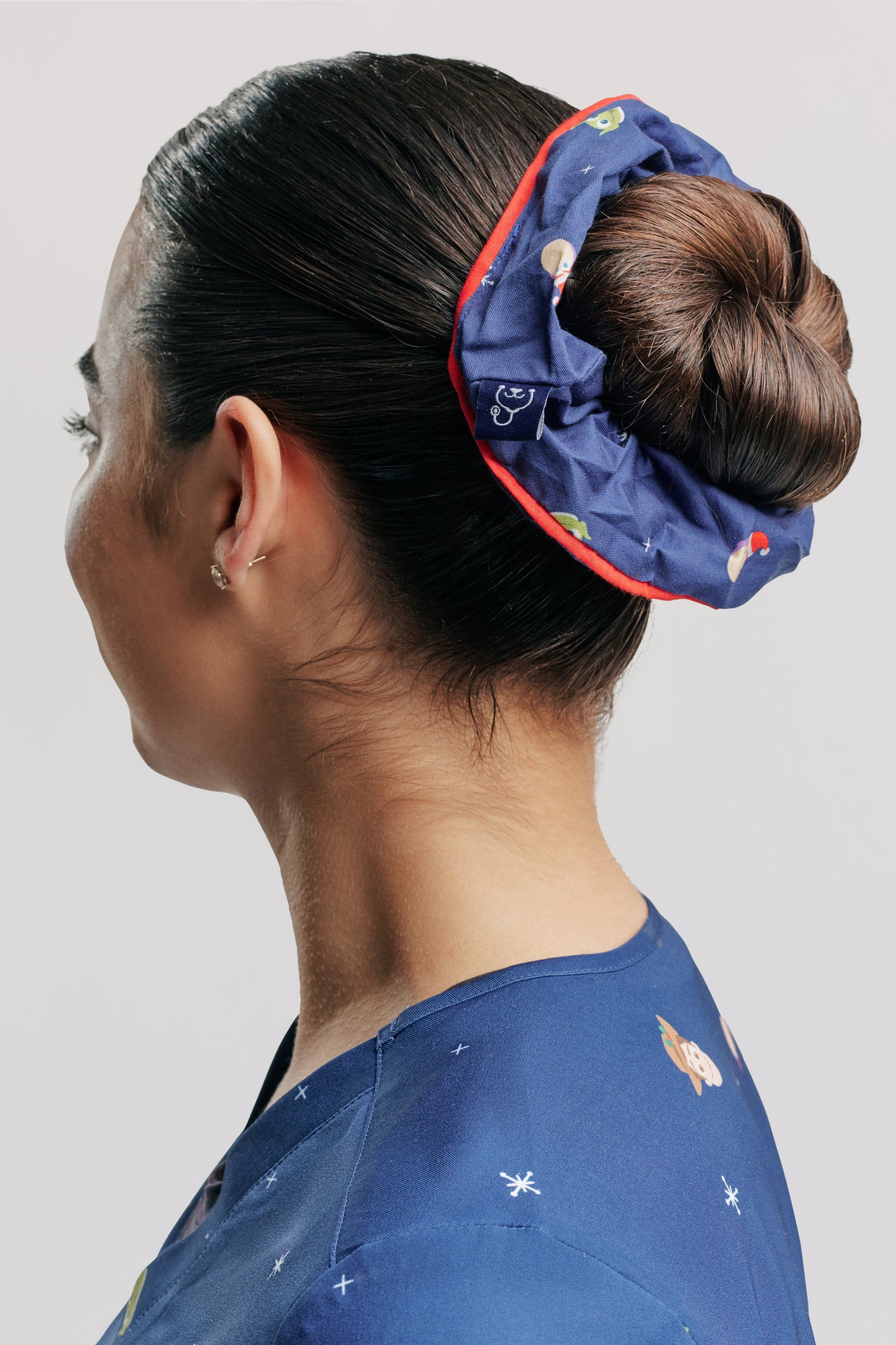 A royal blue Disney & Pixar’s Toy Story Women’s Scrunchie, Ho Ho Howdy in a high bun with red edge piping, paired with the matching blue Toy Story scrub top.   