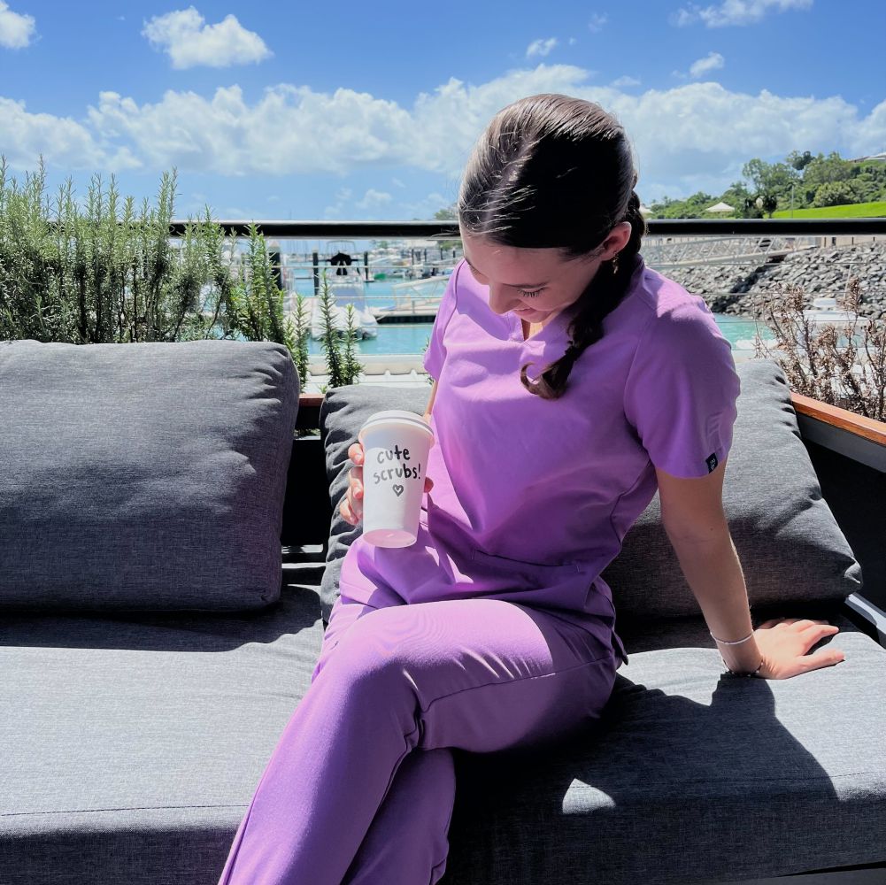 Person in purple scrubs holding cup labeled 'cute scrubs' on a sunny patio.
