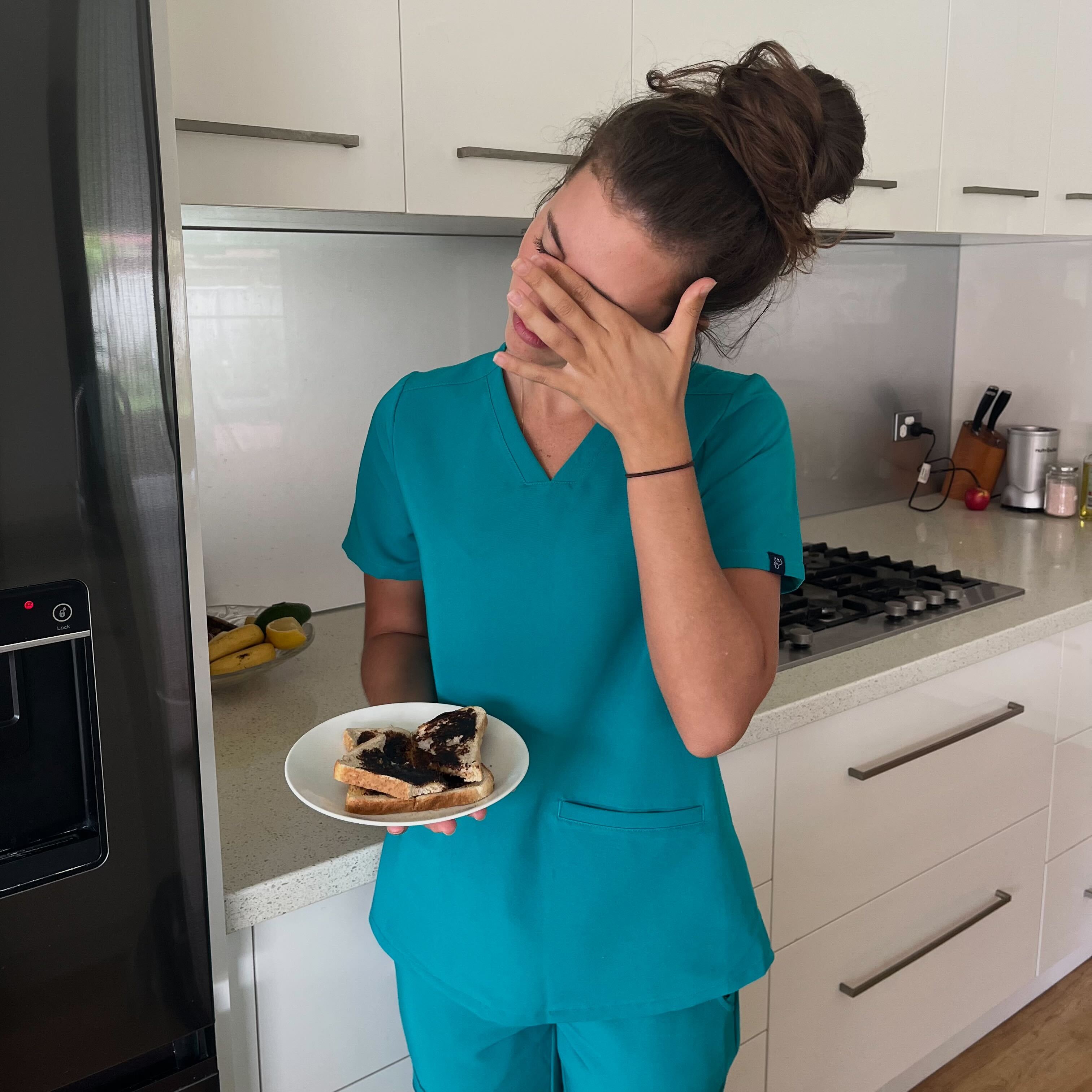 Woman in scrubs holding burnt toast, covering face in disappointment.