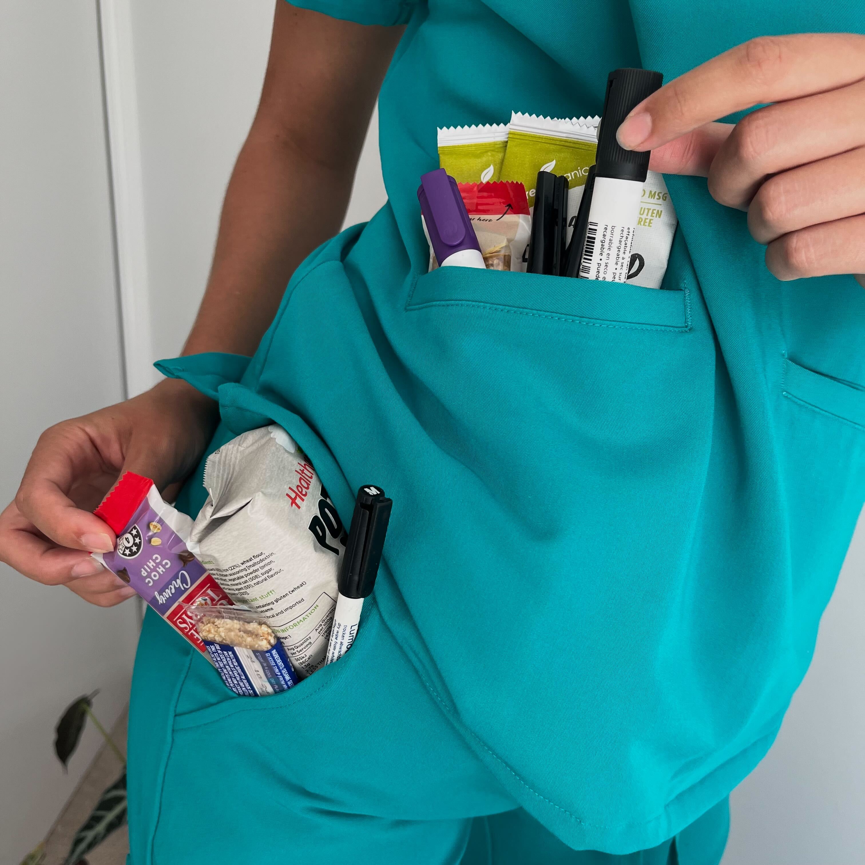 Person in teal scrubs with snacks and markers in pockets.