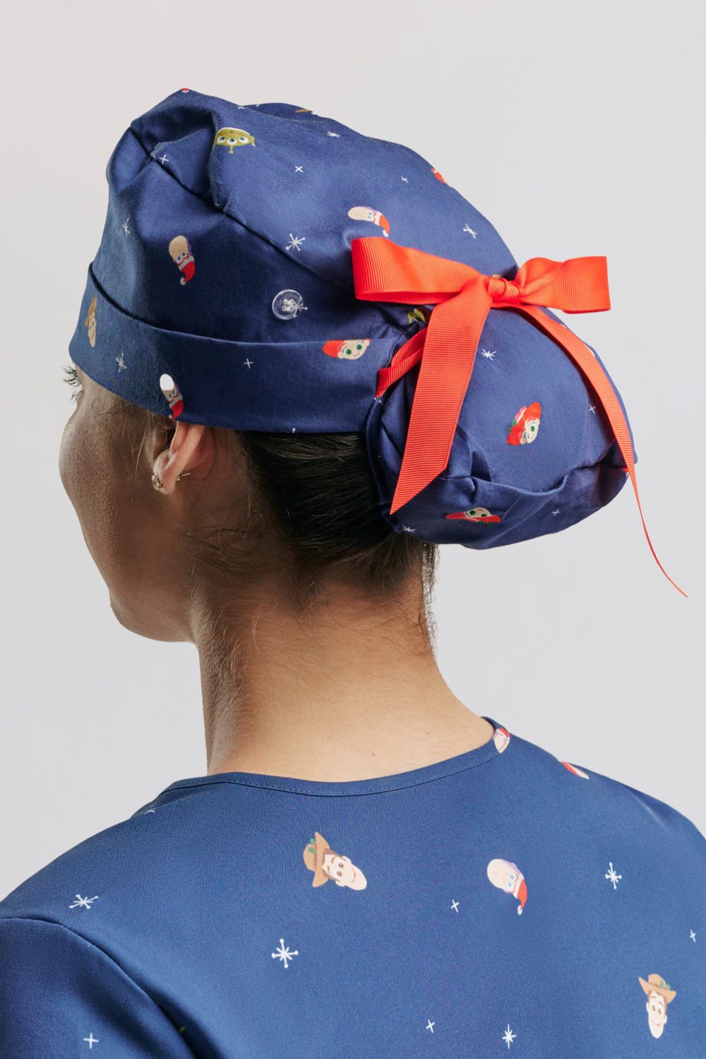 The back profile of the dark blue Disney & Pixar Toy Story Christmas Scrub Cap, highlighting the clear side button for mask straps, the elasticized ponytail pouch secured with a bright red ribbon bow, and the holiday character pattern featuring Woody, Buzz Lightyear in a Santa hat, Jessie, and Squeeze Toy Aliens set against white snowflakes.