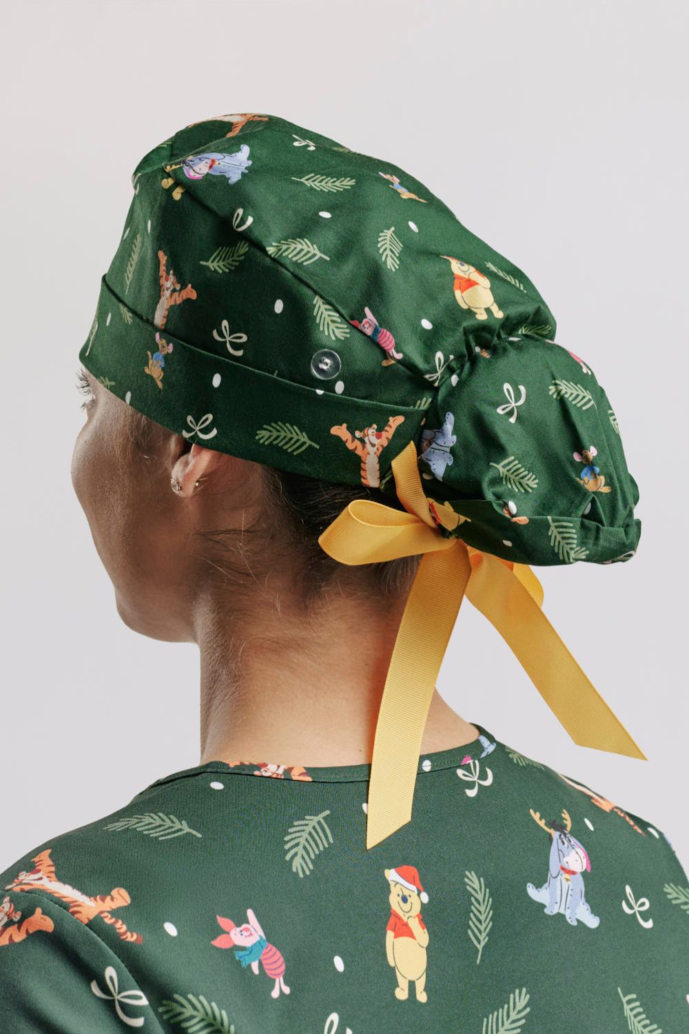 A healthcare worker wearing the dark green Disney’s Winnie the Pooh Christmas Scrub Cap, showcasing the adjustable ponytail pouch design and tied yellow ribbon closure paired with the matching holiday scrub top.   