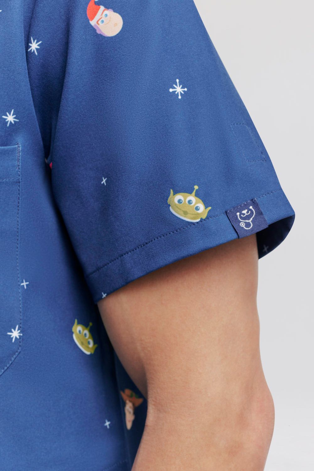 Macro close-up highlighting the sleeve hem and fabric print of the dark blue Disney & Pixar Toy Story Christmas scrub top, featuring illustrations of Buzz Lightyear in a Santa hat, Squeeze Toy Aliens, Woody, and white snowflakes, along with the woven Dr. Woof brand logo tag stitched on the sleeve cuff.