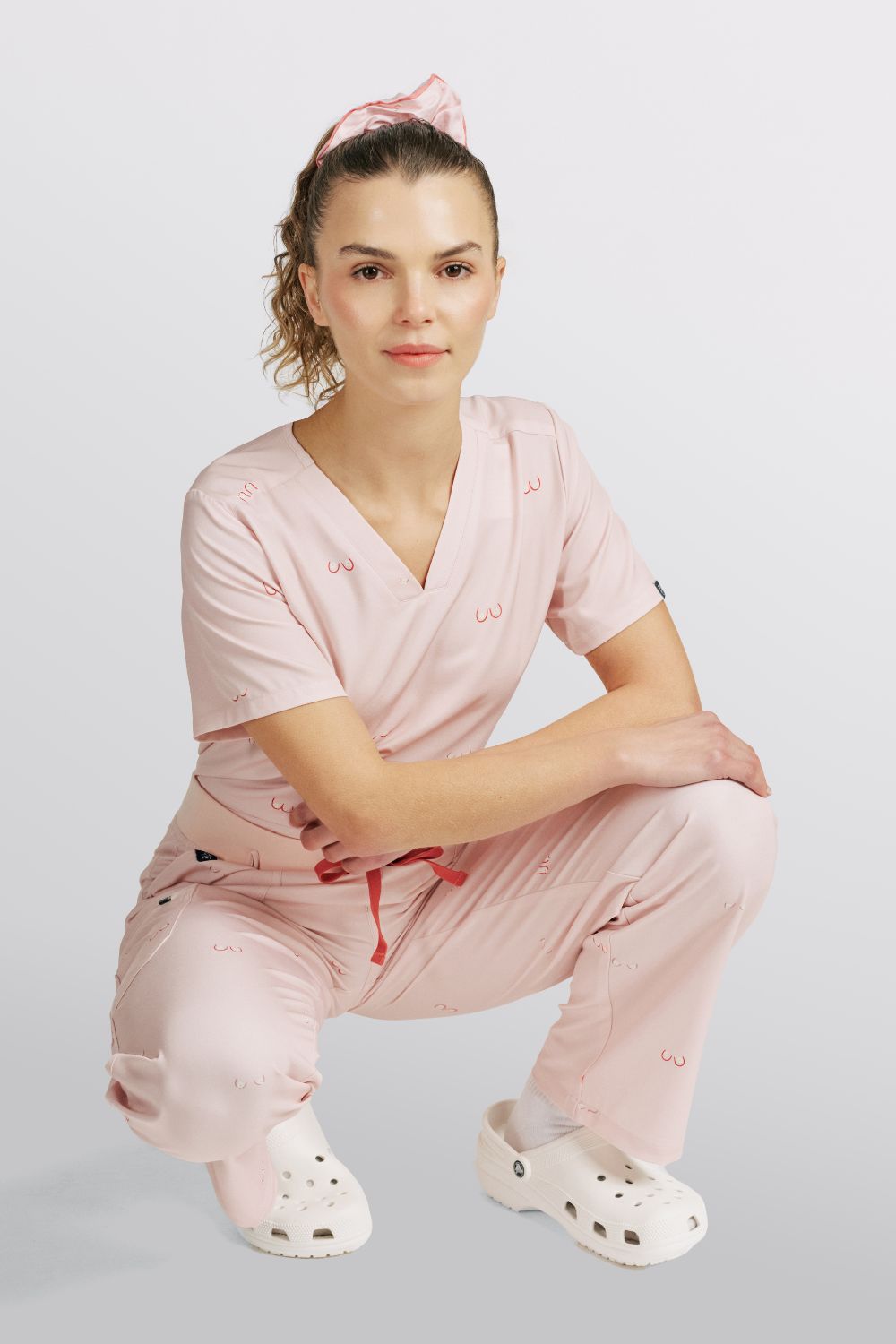 Nurse crouching in light pink scrub top and matching pants with line art boobs print.