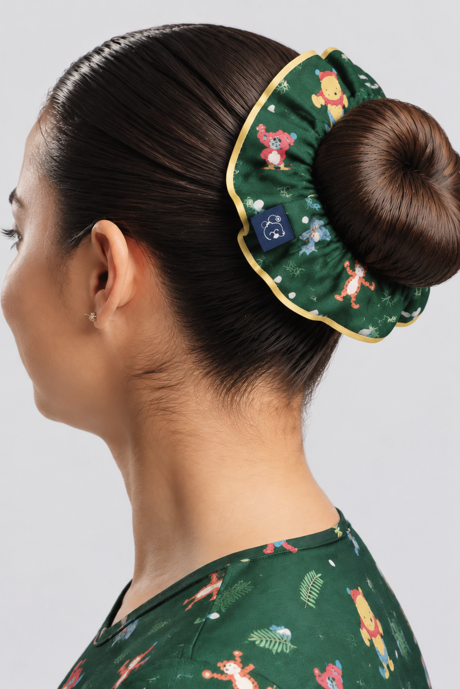 A forest green Disney’s Winnie the Pooh Women’s Scrunchie, Hundred Acre Wood wrapped around a neat hair bun with yellow edge piping, paired with the matching green Hundred Acre Wood scrub top.   