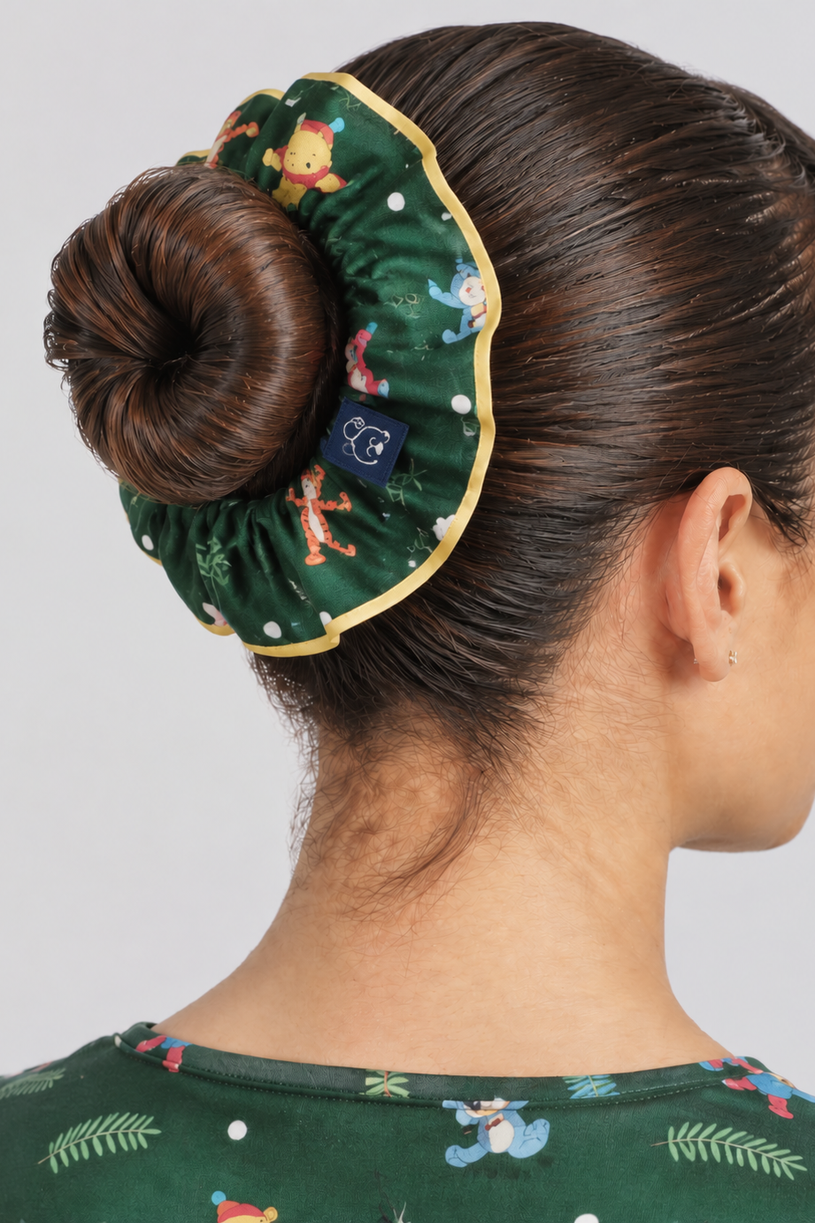 A hair bun with forest green Disney’s Winnie the Pooh Women’s Scrunchie, Hundred Acre Wood wrapped around a neat hair bun with yellow edge piping, paired with the matching green Hundred Acre Wood scrub top.  