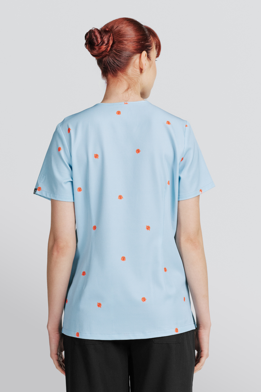 Back view of a nurse wearing a light blue scrub top with a red blood cell pattern and black pants.
